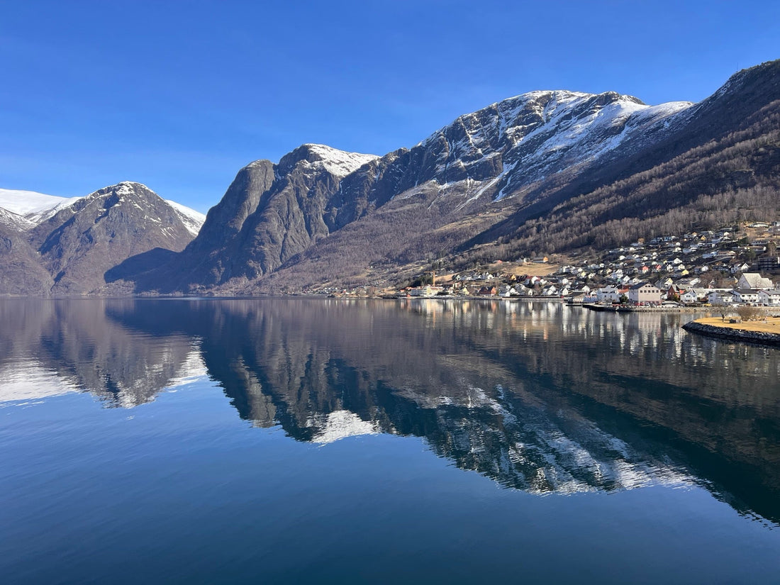 Snow-capped mountains and a calm fjord reflect on a clear blue sky.
